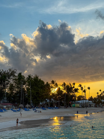 Beach scene with a cloudy sky and a sunset. The sky is filled with clouds, and the sun is setting, creating a beautiful and serene atmosphere. People are enjoying the beachの写真素材