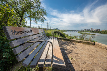 Wooden bench is situated on a path overlooking a river. The bench is empty, and the surrounding area is peaceful and serene. The view of the river is beautifulの写真素材