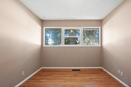 Small, empty room with a white wall and wooden floors. The room has a window with a view of trees...