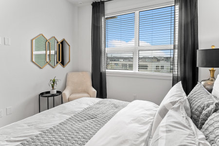 Bedroom with a large window and a white bed with a gray comforter. The room is decorated with a...