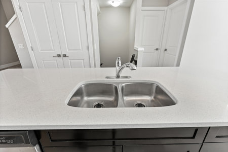 Sink with two basins sits on a white countertop. The sink is stainless steel and has a silver faucet. The countertop is made of granite and is clean and shiny. The kitchen is well-litの写真素材