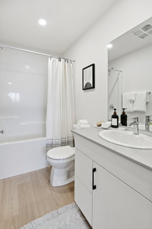Bathroom with a white toilet, sink, and shower. The shower curtain is white and the shower head is...