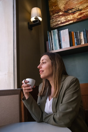 Photo of an entering young woman drinking coffee in a coffee shop smiling and looking out the...