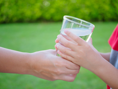Woman hand giving glass of fresh water to child on the green grass backgroundの写真素材