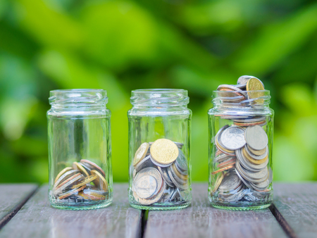 A lot coins in glass money jar on the wood table. Saving for house, health and travel concept.の写真素材