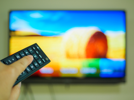 Close up Hand holding TV remote control with a television in the background.の写真素材