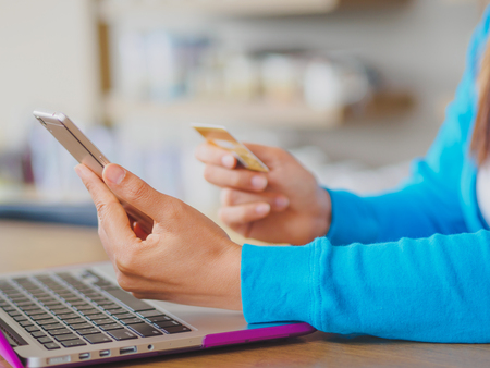 Pretty Young womans hands holding a credit card and using smartphone and laptop computer for online shopping. Online payment. Female working on smart phone laptop in a cafe.の写真素材