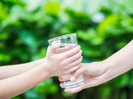 Woman hand giving glass of fresh water to child on the green grass backgroundの写真素材