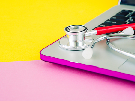 A medical stethoscope and a laptop on a pink and yellow table backgroundの写真素材