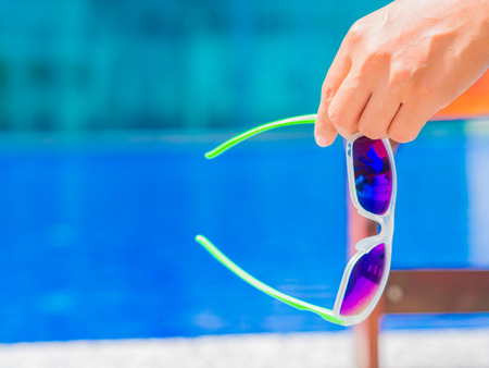 Woman hand holding sunglasses and lying on deckchair by the swimming pool. Vacation and relaxation, summer travel concept.の写真素材