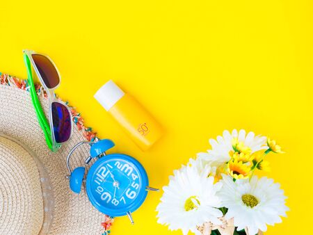 Sunglasses with summer hat, sunscreen lotion, alarm clock and flowers on yellow background.Vacation...