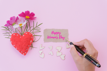 Woman hand holding black pen and writing Happy Women's Day message on brown paper with handmade red heart, beautiful flowers, leaf and a lot of wooden hearts on pink background. Womens Day concept.の写真素材