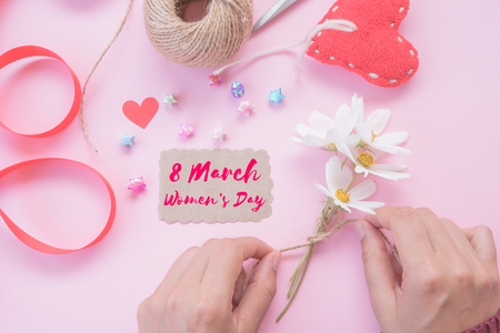 Woman hand holding white flowers with writing Happy International Women's Day message on white paper with handmade red heart, beautiful flowers, leaf, red ribbon, colorful of paper stars and a lot of wooden hearts on pink background. Womens Day concept.の写真素材