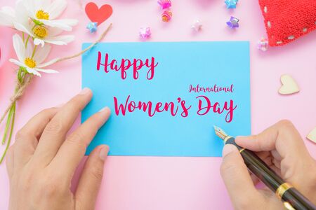 Woman hand holding black pen and writing Happy International Women's Day message on blue paper with handmade red heart, beautiful flowers, colorful of paper stars and wooden hearts on pink background. Womens Day concept.の写真素材