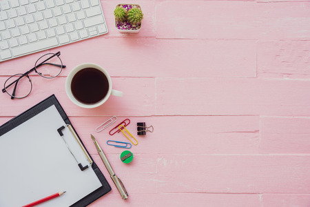 Top view office table with workspace and office accessories including keyboard, glasses, clips, flower, pen, note book, Clipping document and coffee on pink wooden background.の写真素材