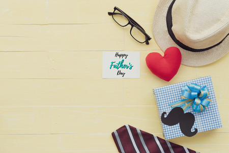 Happy fathers day concept. Red tie, glasses, hat, mustache, gift box with Happy father's day text and handmade red heart on bright yellow pastel wooden table background.の写真素材