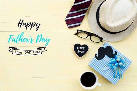 Happy fathers day concept. Red tie, glasses, hat, mustache, gift box with Love DAD text on black heart tag on bright yellow pastel wooden table background.の写真素材