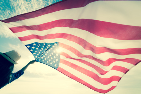 USA Independence day, 4 July. Close up oung happy woman holding United States of America flag and running, jumping carefree with open arms with blue sky.の写真素材