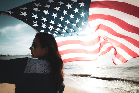 USA Independence day, 4 July. Close up oung happy woman holding United States of America flag and running, jumping carefree with open arms on the beach.の写真素材