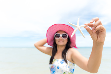 Beautiful woman wearing hat beach and sunglasses and holding starfish on hands over sandy beach, green sea and blue sky background for summer holiday and vacation concept.の写真素材