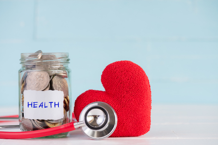 Money saving and health care concept. A jar contains coins with stethoscope and handmade red heart on white and pastel green wooden background.の写真素材