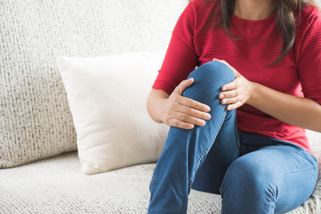 Closeup young woman sitting on sofa and feeling knee pain and she massage her knee at home. Healthcare and medical concept.の写真素材