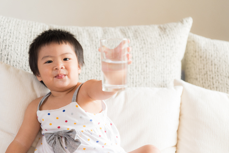 Happy little asian girl hand holding drinking water glass and laughing, sitting on sofa at home. Medicine and health care concept.の写真素材