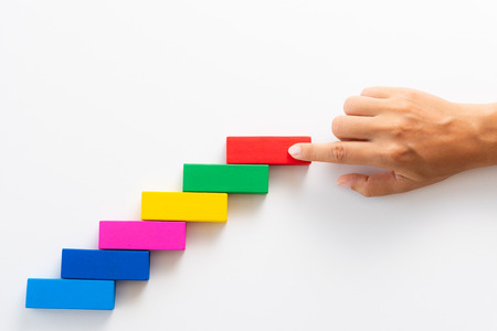 Concept of building success foundation. Women hand put red wooden block on colorful wooden blocks in the shape of a staircase.の写真素材