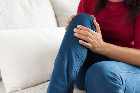 Closeup young woman sitting on sofa and feeling knee pain and she massage her knee at home. Healthcare and medical concept.の写真素材