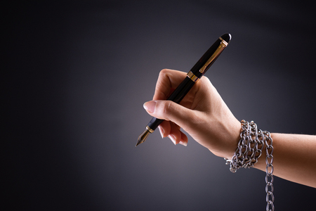 Woman hand with black pen tied with chain, depicting the idea of freedom of the press or freedom of expression on dark background in low key. World press freedom and international human rights day concept.の写真素材