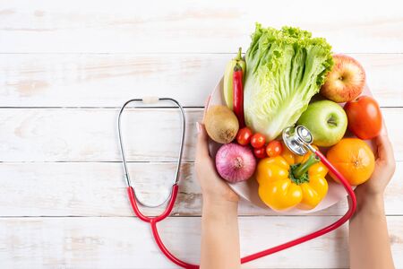 Healthy lifestyle, food and nutrition concept. Close up doctor woman hand holding plate of fresh vegetables and fruits with stethoscope lying on white wooden table.の写真素材