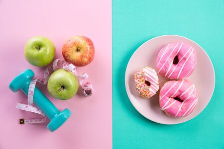 Healthy lifestyle, food and sport concept. Top view of healthy versus unhealthy. donut versus measuring tape with athlete's equipment dumbbell, green red apple on pink blue pastel background.の写真素材