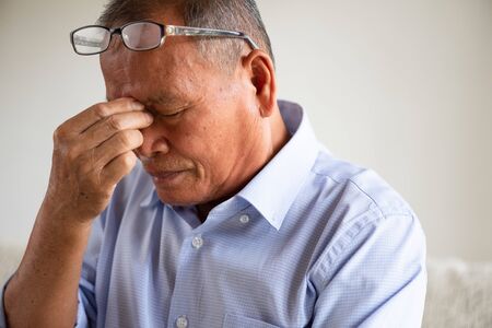 Asian old man sitting on sofa and having a headache at home. Senior healthcare concept.の写真素材