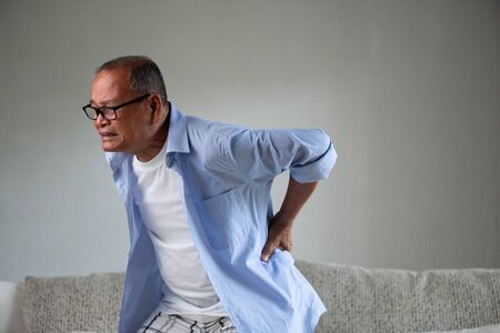 Asian old man sitting on sofa and having a back pain, backache at home. Senior healthcare concept.の写真素材