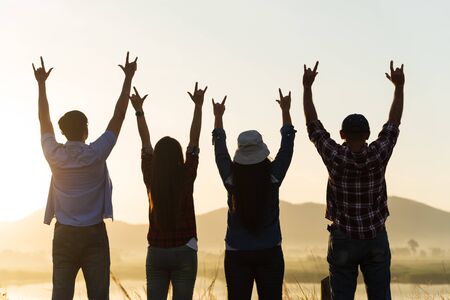 Group of happy friends are having fun with raised arms together in front of mountain and enjoy sunrise sunset showing unity and teamwork. Friendship happiness leisure partnership team concept.の写真素材
