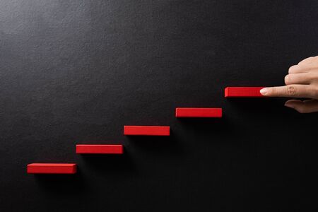 Concept of building success foundation. Women hand put red wooden block in the shape of a staircase....
