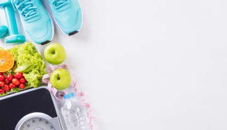 Healthy lifestyle, food and sport concept. Top view of sport shoes, weight scale measuring tape, blue dumbbell, sport water bottles, fruit and vegetables on white wooden background.の写真素材