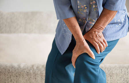 Asian old woman sitting on sofa and having knee pain, knee injury at home. Senior healthcare concept.の写真素材