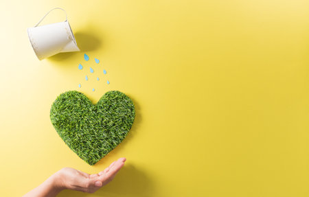 Hands holding green heart with watering on yellow background. World environment day, earth day, save earth and eco concept.の写真素材
