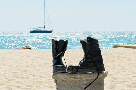 Pair of boots on the sand beach with water and boat on the foregroundの写真素材