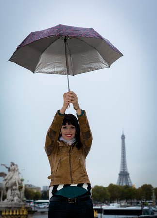 Young smiling brunette on vacation in Paris France holding umbrella in her hands. Eiffel tower is visible on background.の写真素材