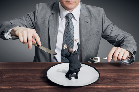 Businessman holding fork with knife and ready to eat small scared businessman on the plate. Hungry businessman ready to eating a competitorの写真素材