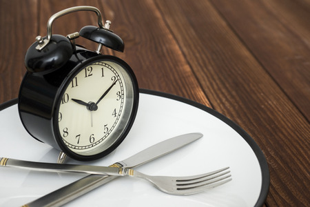 Alarm clock on plate with knife and fork on wooden background. Time to eat. Weight loss or diet...