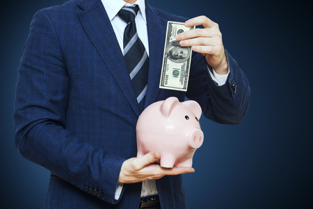 Businessman putting one hundred dollar into piggy bank. Businessman in suit is holding piggy bank. Finance Savings conceptの写真素材