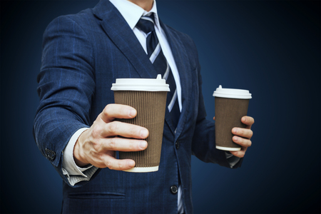 Businessman offering coffee in paper cup. Businessman offers a cup of coffee. Businessman holding two disposable coffee cupの写真素材