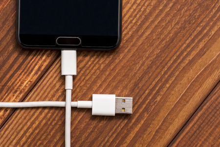 Smartphone with USB cable on the wooden table. Smartphone or tablet with USB cable prepared for data transfer or battery chargingの写真素材