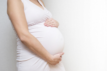 Pregnant woman in dress holds hands on belly on light background. Pregnancy, maternity, preparation and expectation concept. Motherhood, pregnancy, happiness concept, copy spaceの写真素材