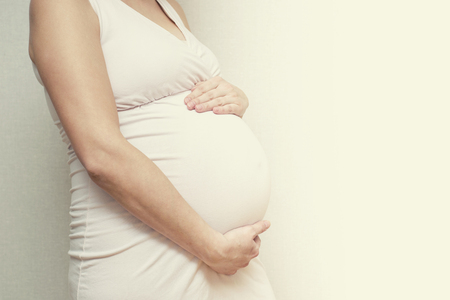 Pregnant woman in dress holds hands on belly on light background. Pregnancy, maternity, preparation and expectation concept. Motherhood, pregnancy, happiness concept, copy spaceの写真素材
