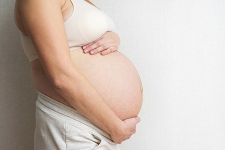 Pregnant woman holds hands on belly on light background. Pregnancy, maternity, preparation and expectation concept. Close-up, copy space, indoors. Beautiful tender mood photo of pregnancyの写真素材