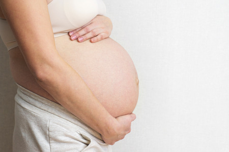 Pregnant woman holds hands on belly on light background. Pregnancy, maternity, preparation and expectation concept. Close-up, copy space, indoors. Beautiful tender mood photo of pregnancyの写真素材